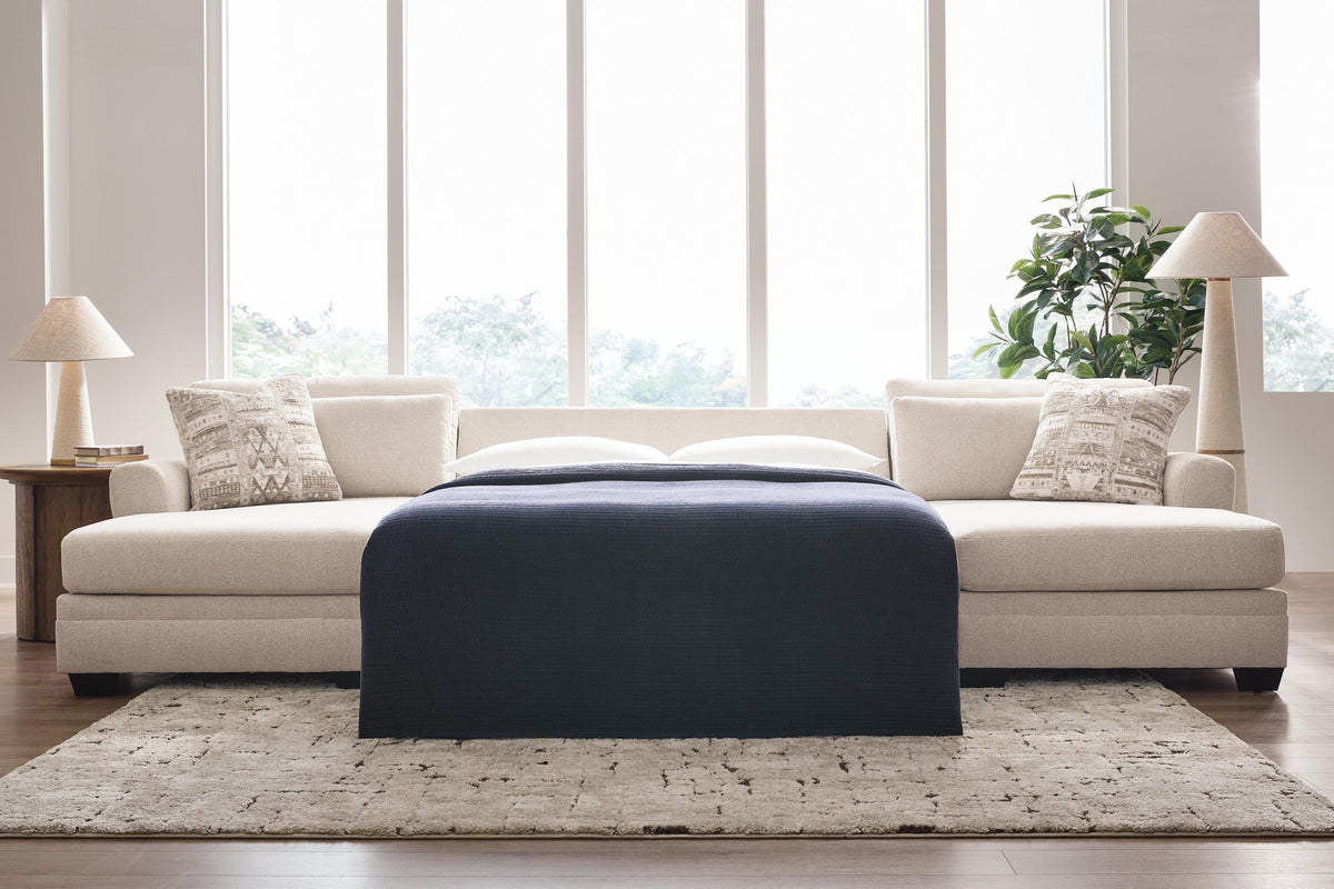 A modern sectional sofa in cream with patterned accent pillows and a dark navy throw blanket, styled in a bright living room with a textured rug, side tables, lamps, and a plant.