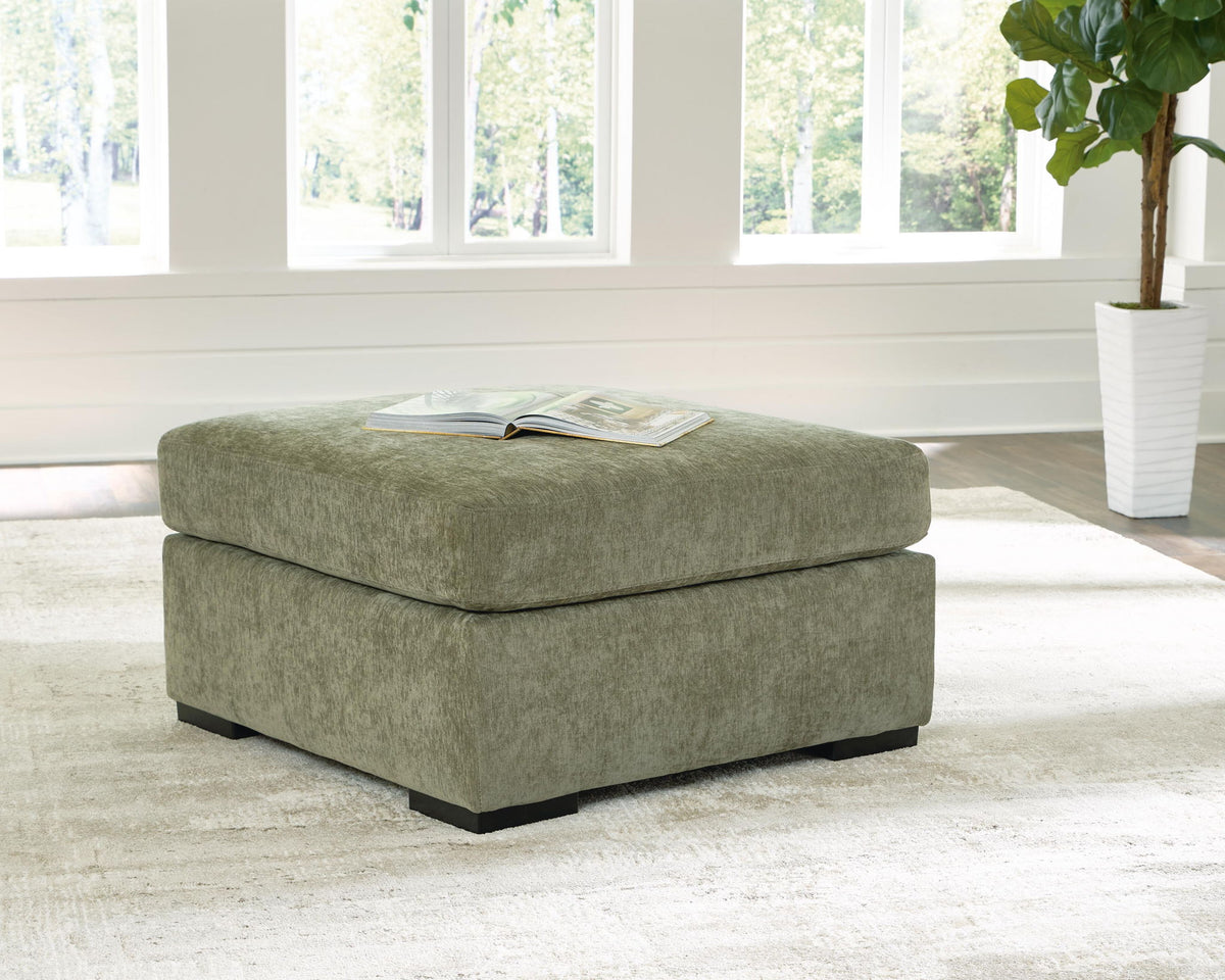 A square upholstered ottoman in textured sage green fabric, shown in a bright living room setting with an open magazine resting on top, set against a white shiplap wall and large windows.
