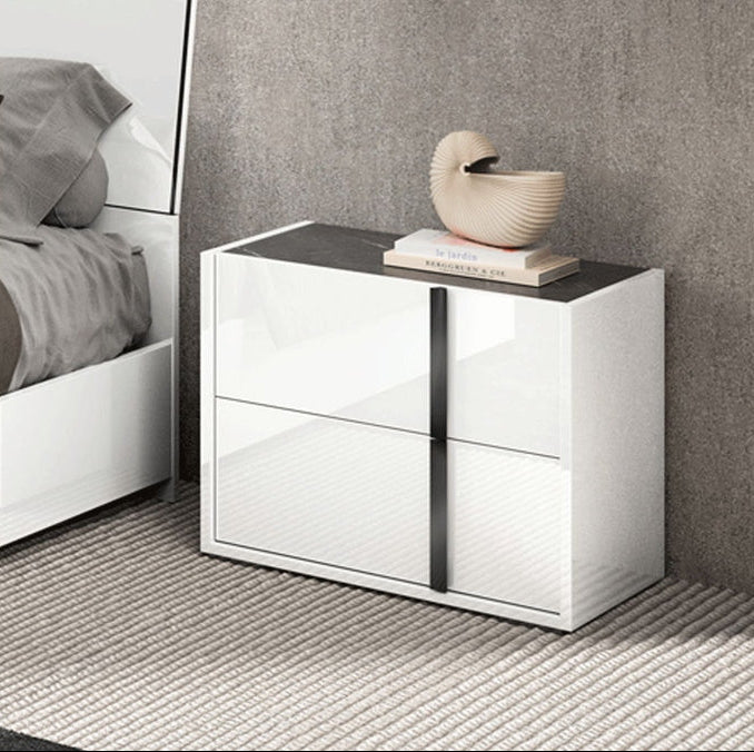 A modern two-drawer nightstand with a glossy white finish and a dark top surface, styled in a bedroom setting next to a bed.