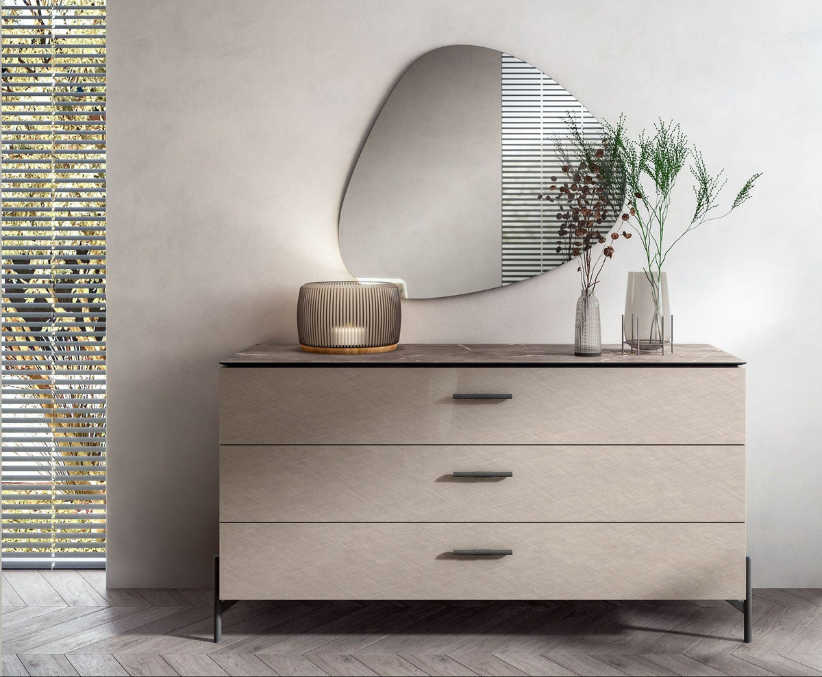 A modern three-drawer dresser with a light beige textured finish and dark gray top, styled in a minimalist room with an organic-shaped mirror, a small lamp, and two glass vases on top.