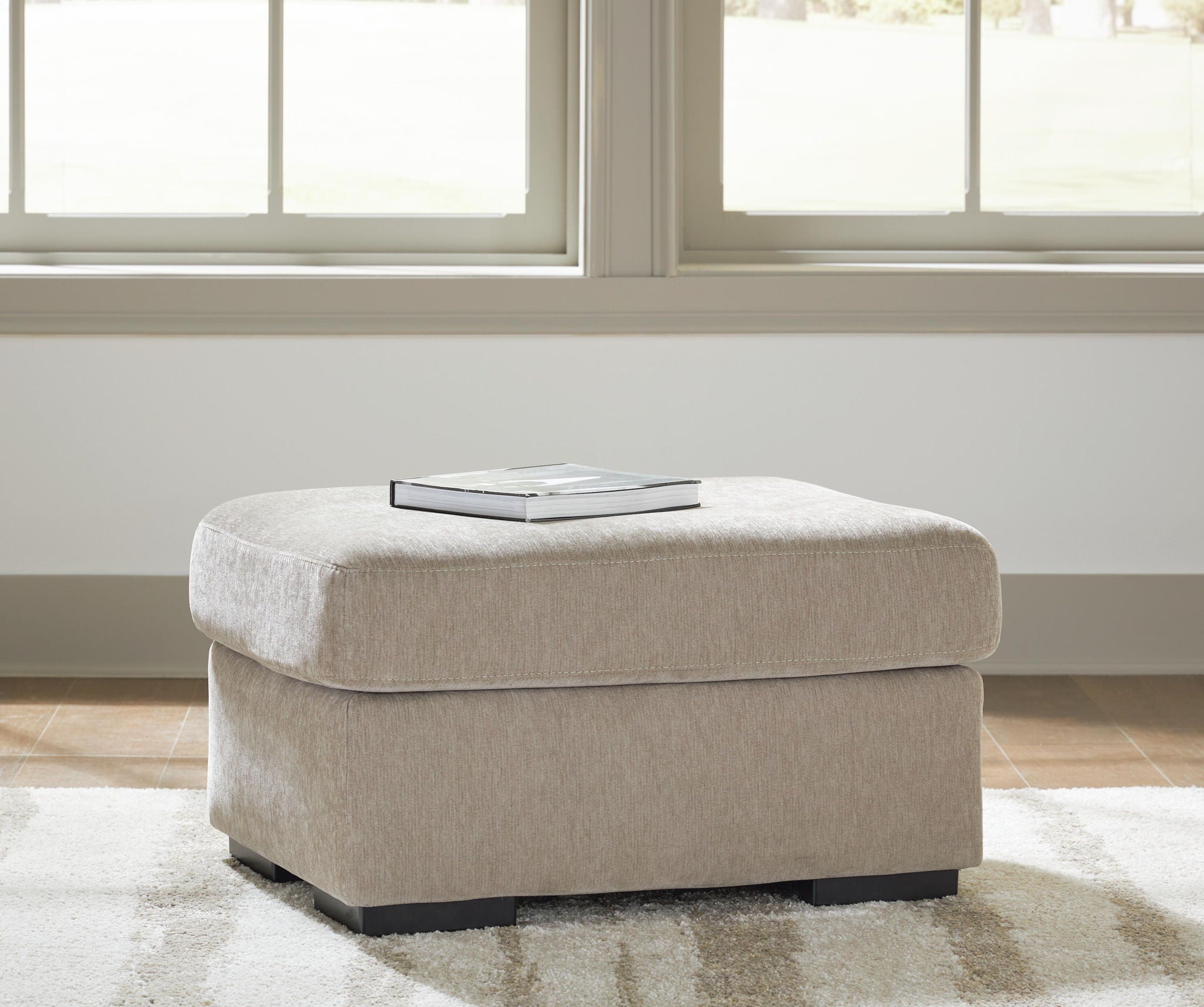 A square upholstered ottoman in light beige fabric sits on a patterned rug in front of a bright window. A book rests on top of the ottoman, and the room has light wood flooring and white walls.