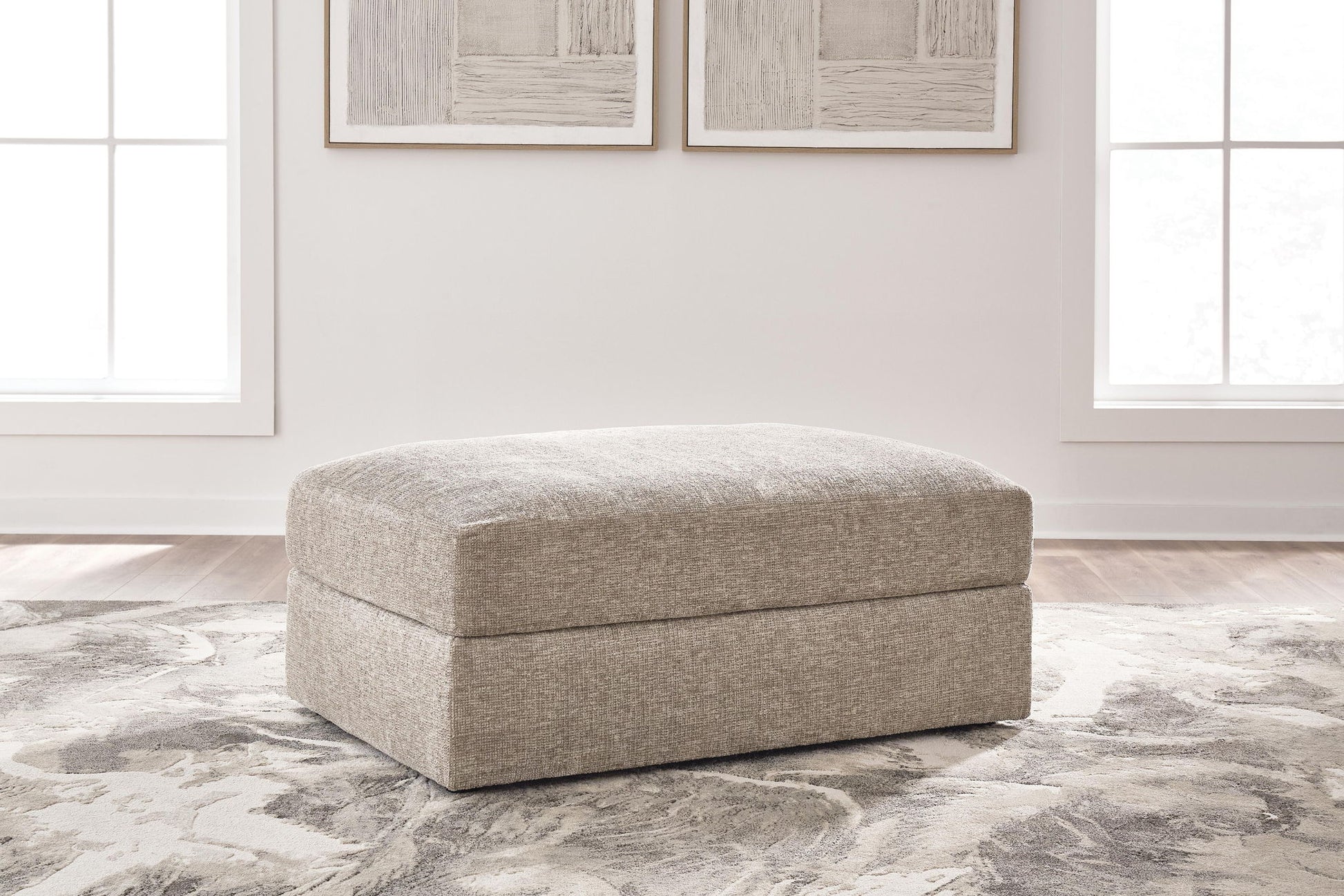A square upholstered ottoman in a textured light beige fabric, positioned on a gray and white patterned area rug in a bright, modern room with two windows and framed wall art.