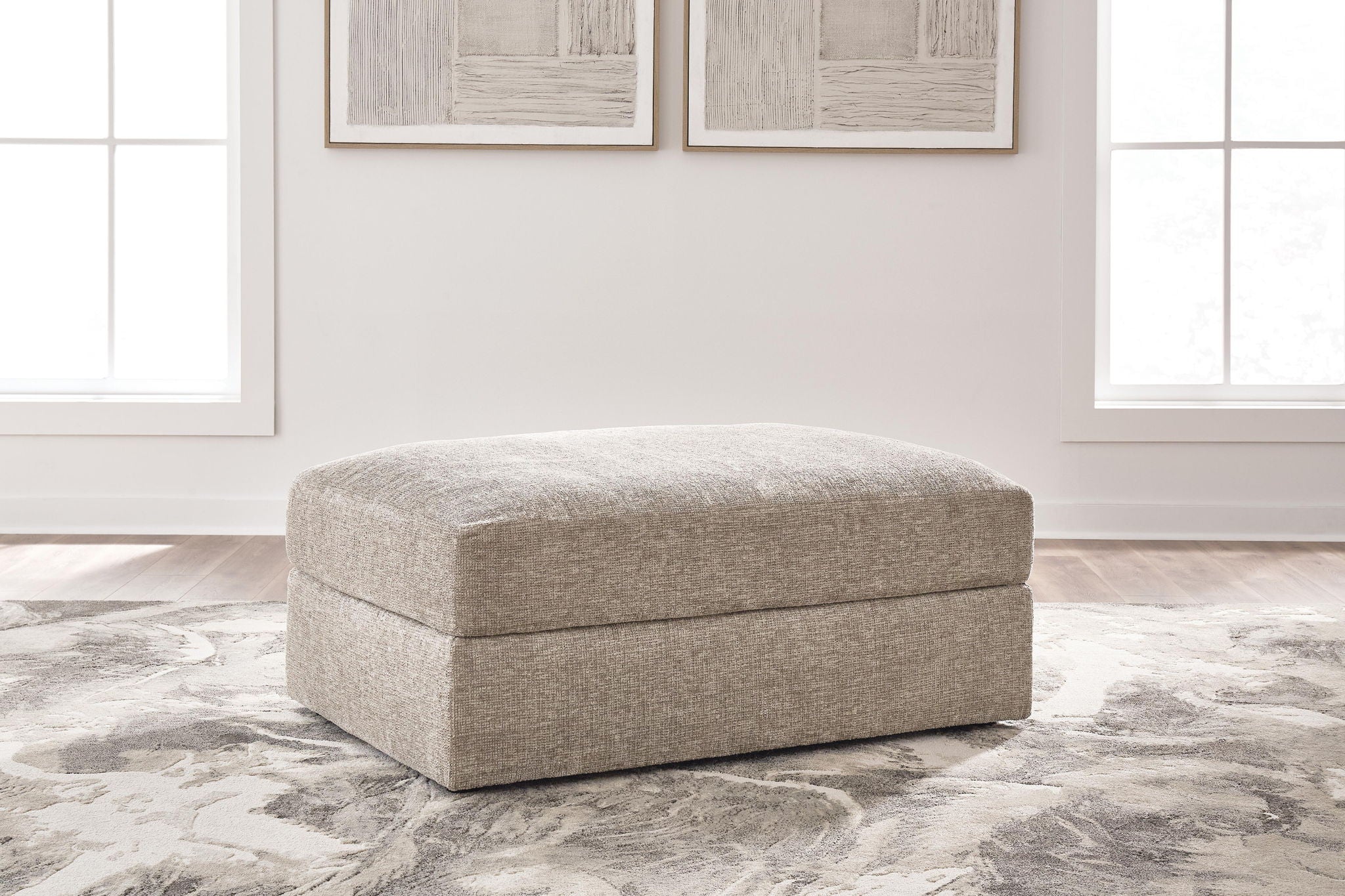 A square upholstered ottoman in a textured light beige fabric, positioned on a gray and white patterned area rug in a bright, modern room with two windows and framed wall art.
