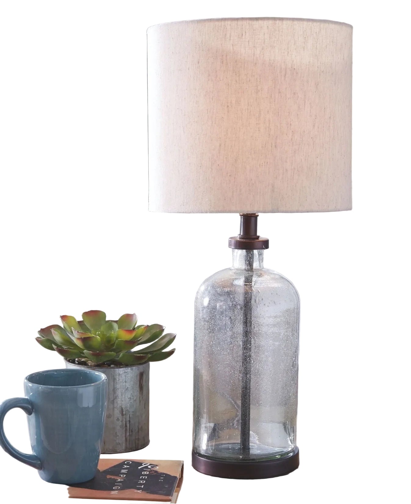 Glass table lamp with a beige shade on a wooden surface, next to a blue mug and small plant.