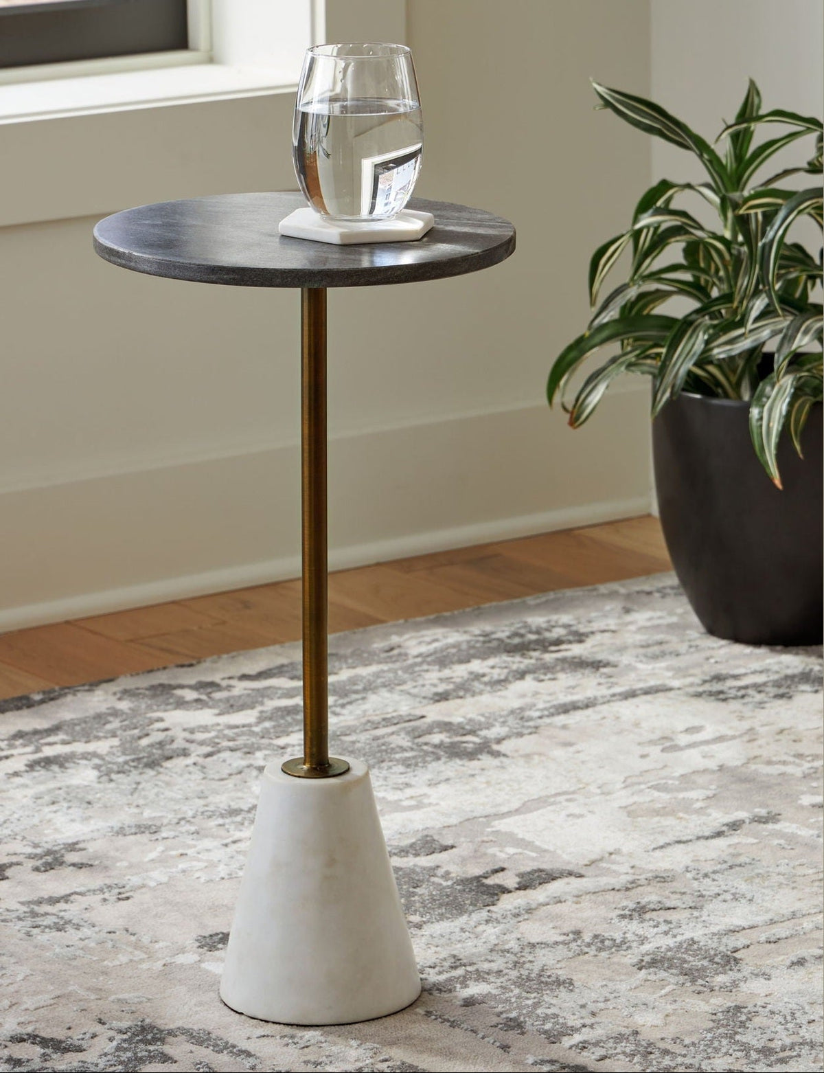 Small round side table with a white base and gold leg in a room with a plant and window.