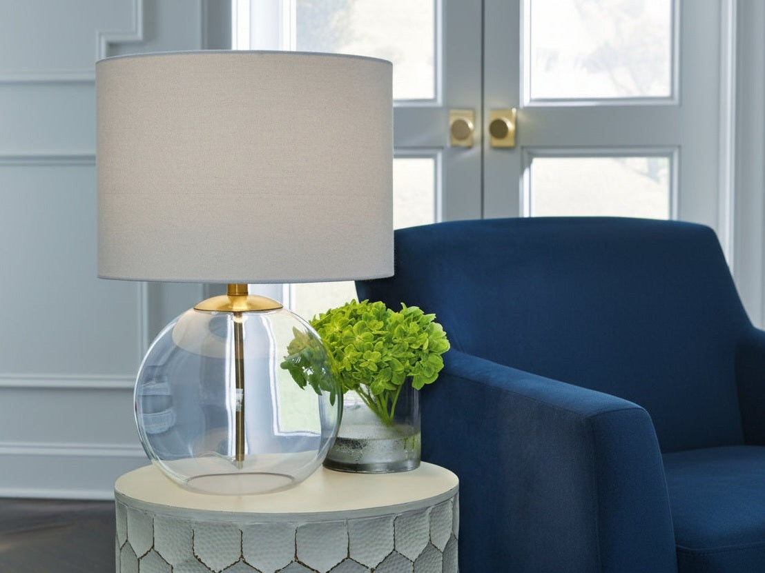 Samder table lamp with a clear glass base and beige shade on a side table next to a blue chair.