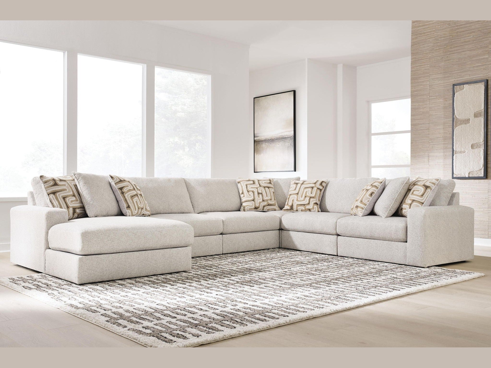 A light beige sectional sofa with a chaise lounge on the left, styled with patterned throw pillows, set on a textured gray and white area rug in a bright, modern living room.