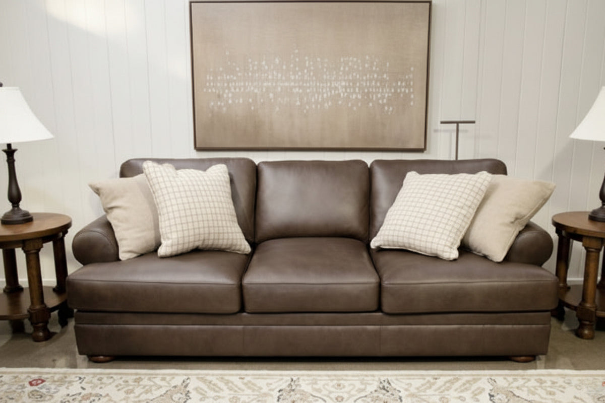 A three-seater brown leather sofa with four cream throw pillows, set in a living room environment with a patterned rug, side tables, and wall art.