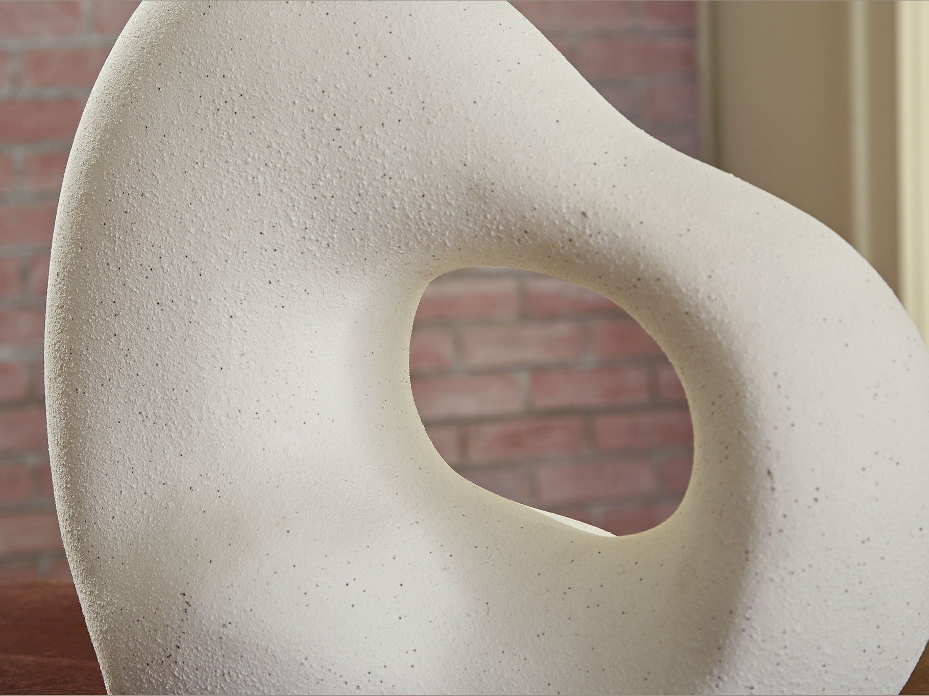Abstract stone sculpture with a circular hole against a brick wall.