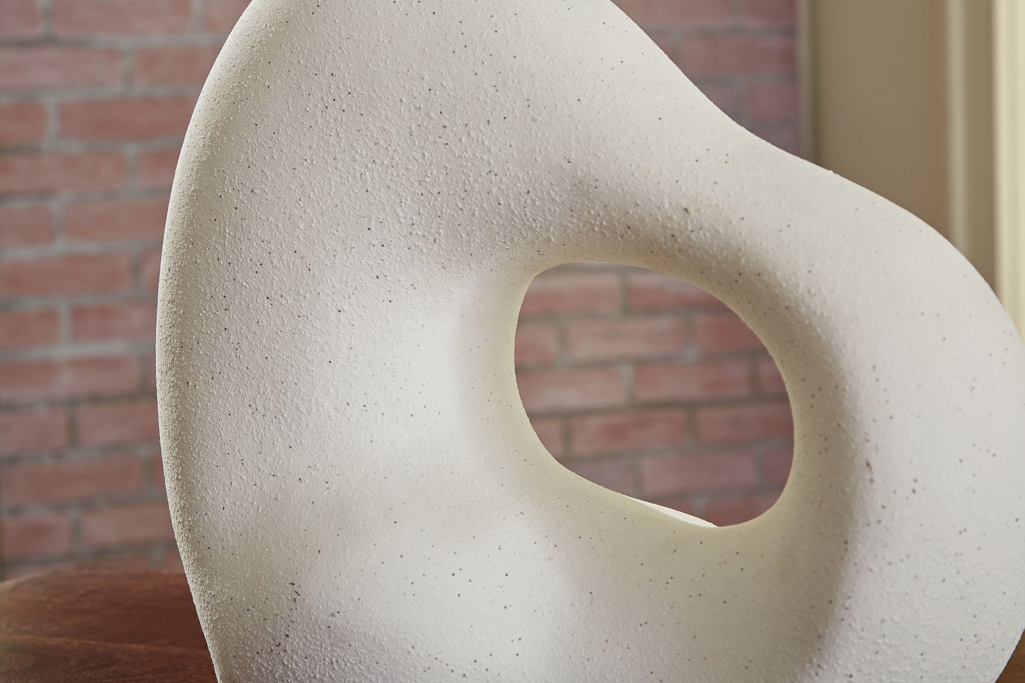 Abstract stone sculpture with a circular hole against a brick wall.