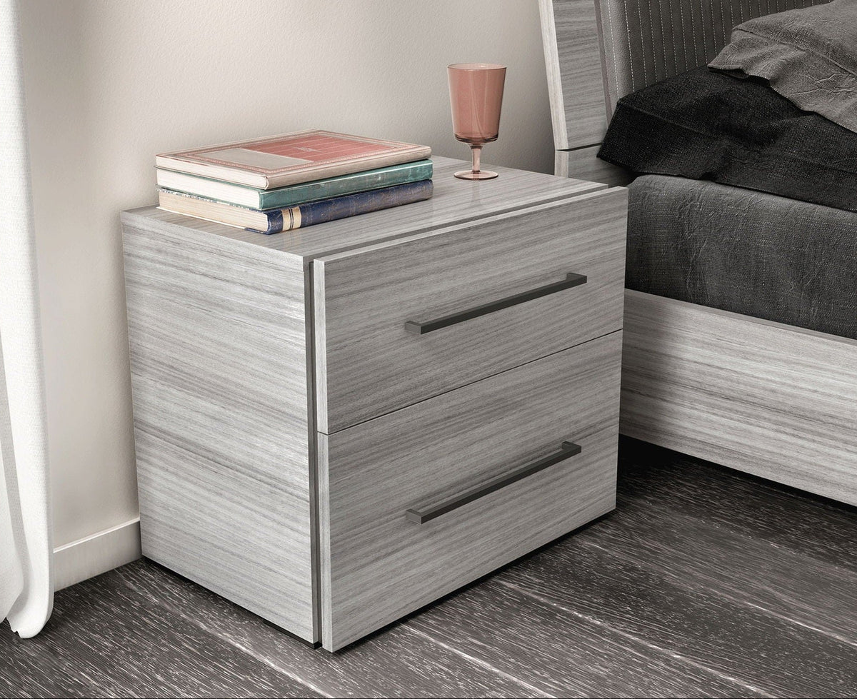 A two-drawer nightstand with a light gray wood-grain finish and dark metal handles, styled in a bedroom next to a bed with gray bedding.