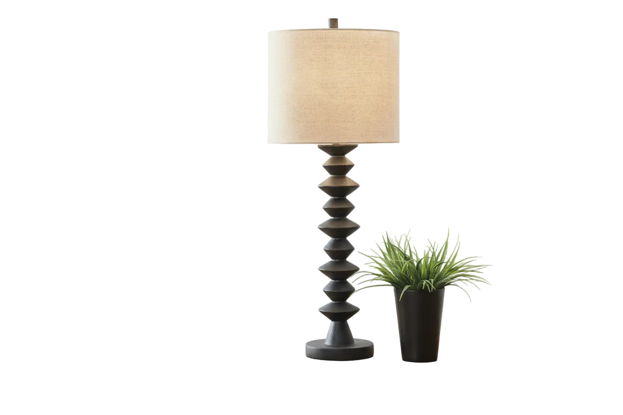 Floor lamp with a textured base on a wooden table in a room with a couch and decorative plant.