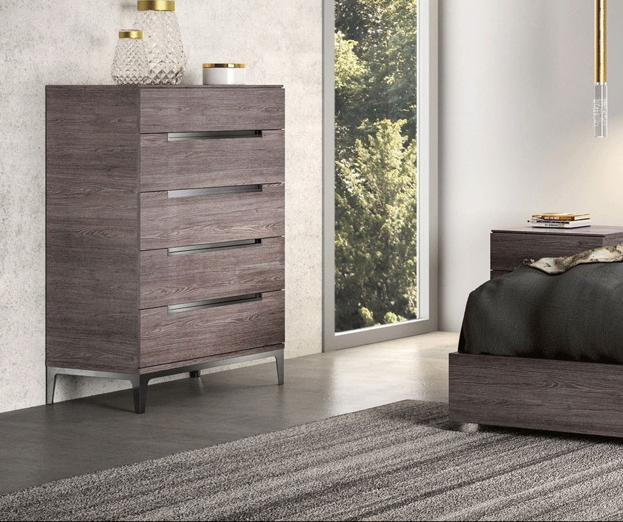 A modern four-drawer chest of drawers in a dark brown wood grain finish with slim horizontal metal handles, shown in a styled bedroom setting next to a matching bed.