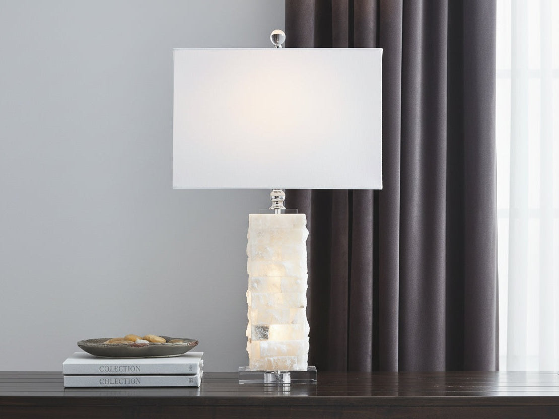 Malise table lamp with a textured white base and white shade against a gray wall and dark curtains.