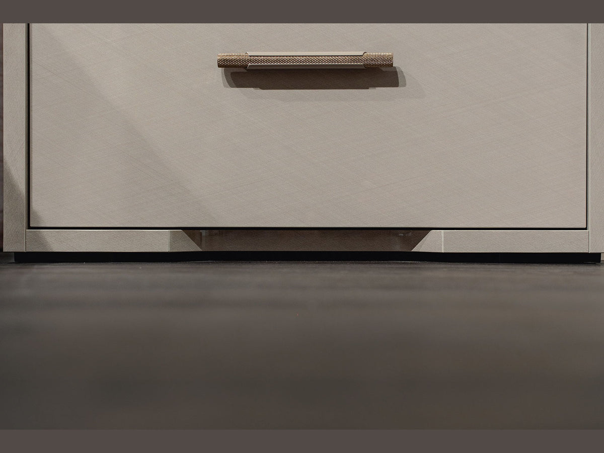 Close-up of a modern drawer front in a warm light beige tone with a slim horizontal metallic handle, set against a dark gray floor.