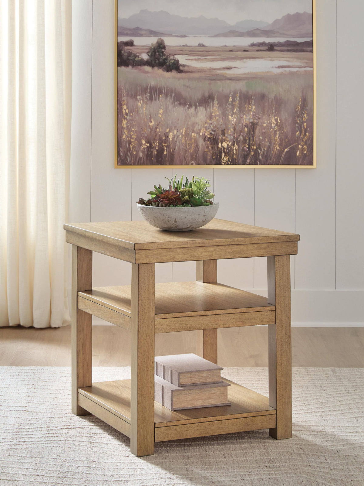 A two-tier wooden side table in a light natural oak finish, styled with a small bowl of succulents on top and books on the lower shelf, set in a bright room with a framed landscape painting on the wall.