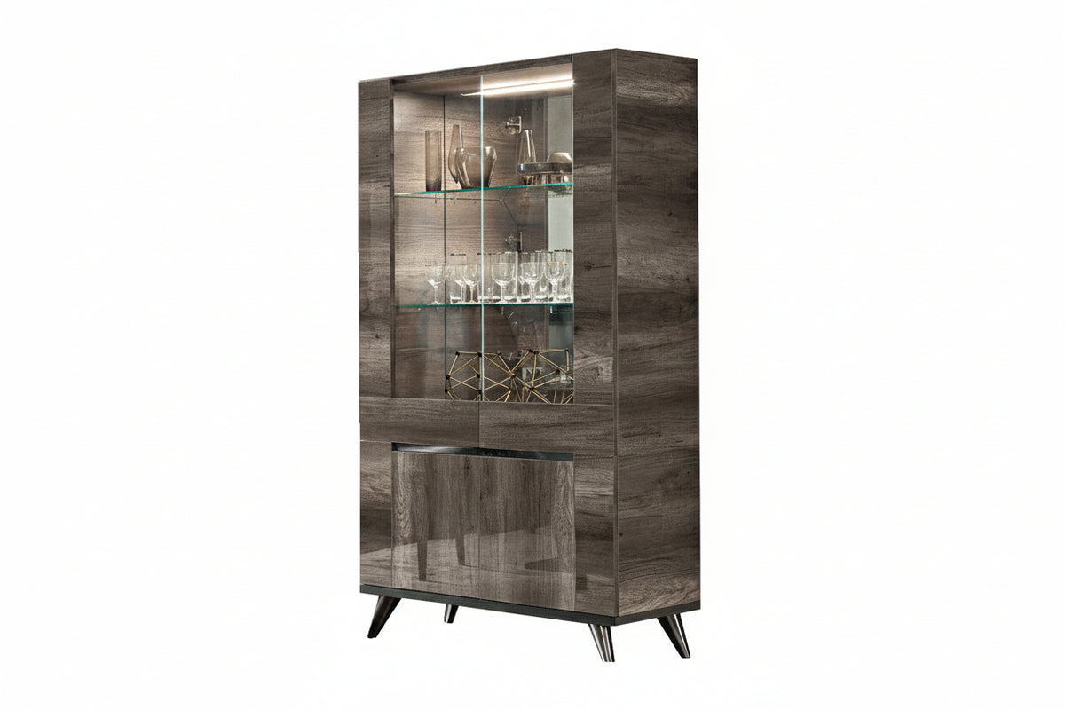 A tall modern glass-front display cabinet with a gray wood-grain finish, featuring three glass shelves with wine glasses and decorative items, illuminated interior lighting, and tapered black legs, shown on a plain white background.