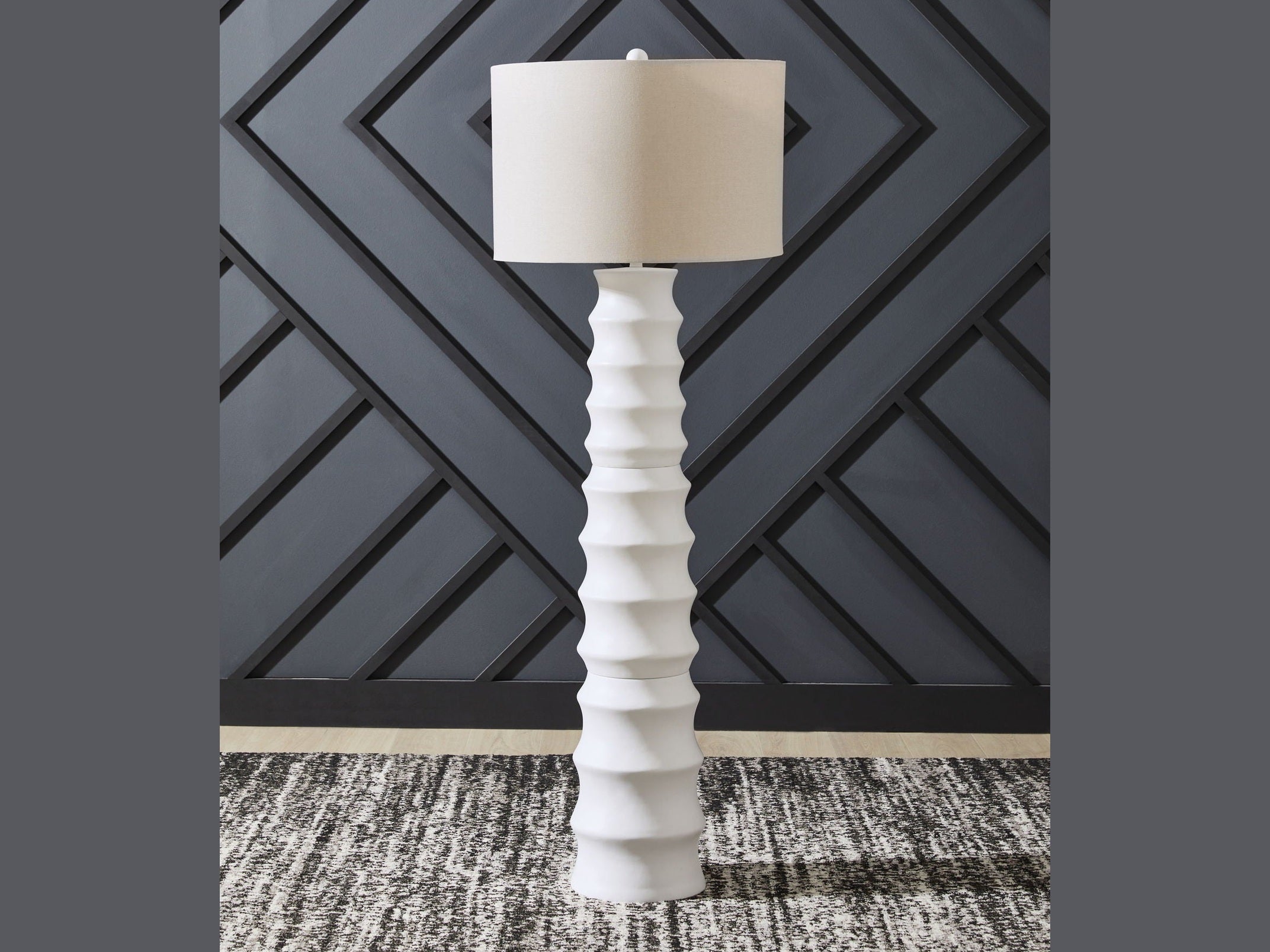 White floor lamp with a beige shade against a geometric-patterned wall.