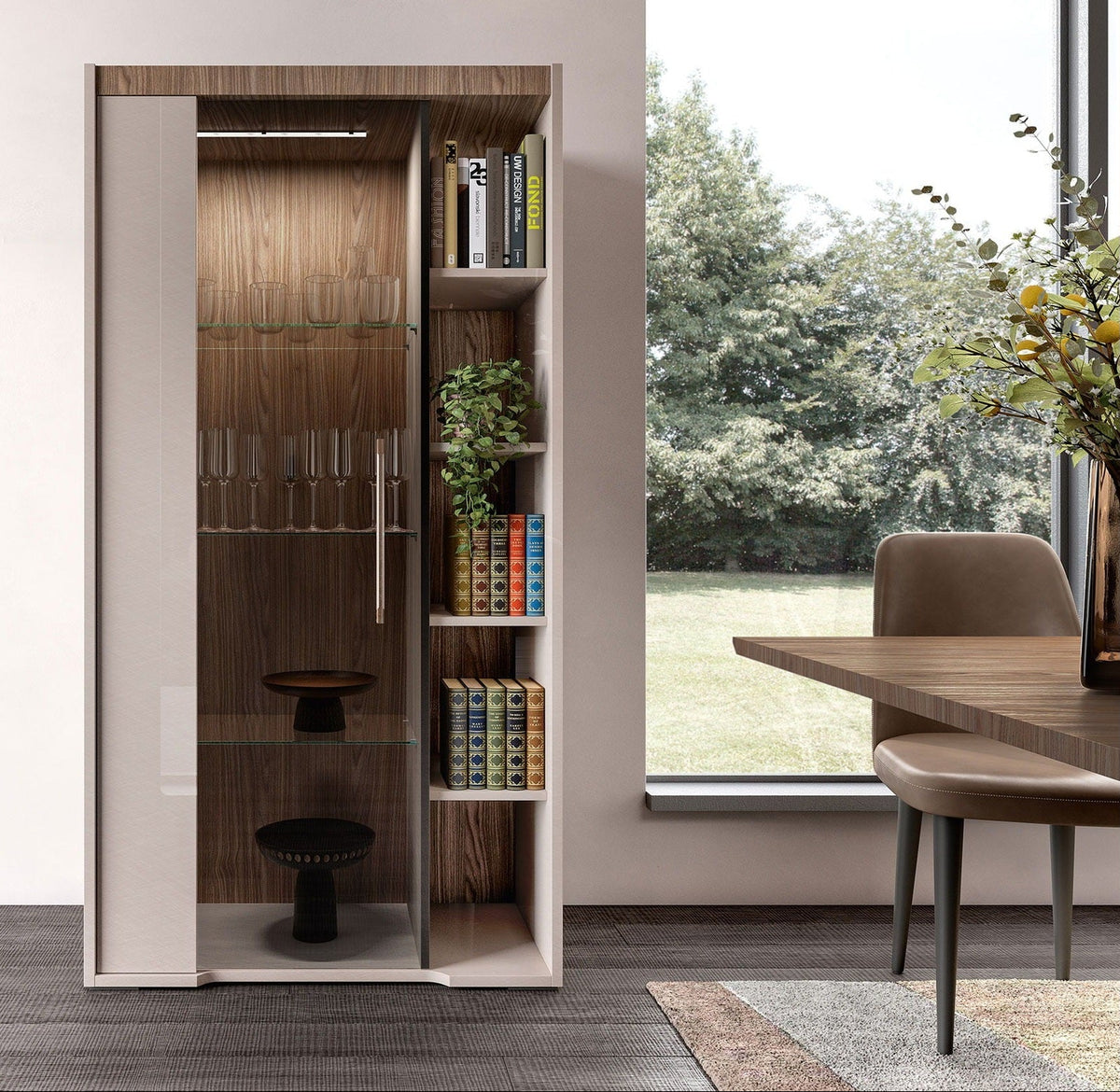 A modern tall storage cabinet with sliding beige doors and a wood-grain interior, shown in a styled room next to a window with a view of trees, with a dining table and chair nearby.
