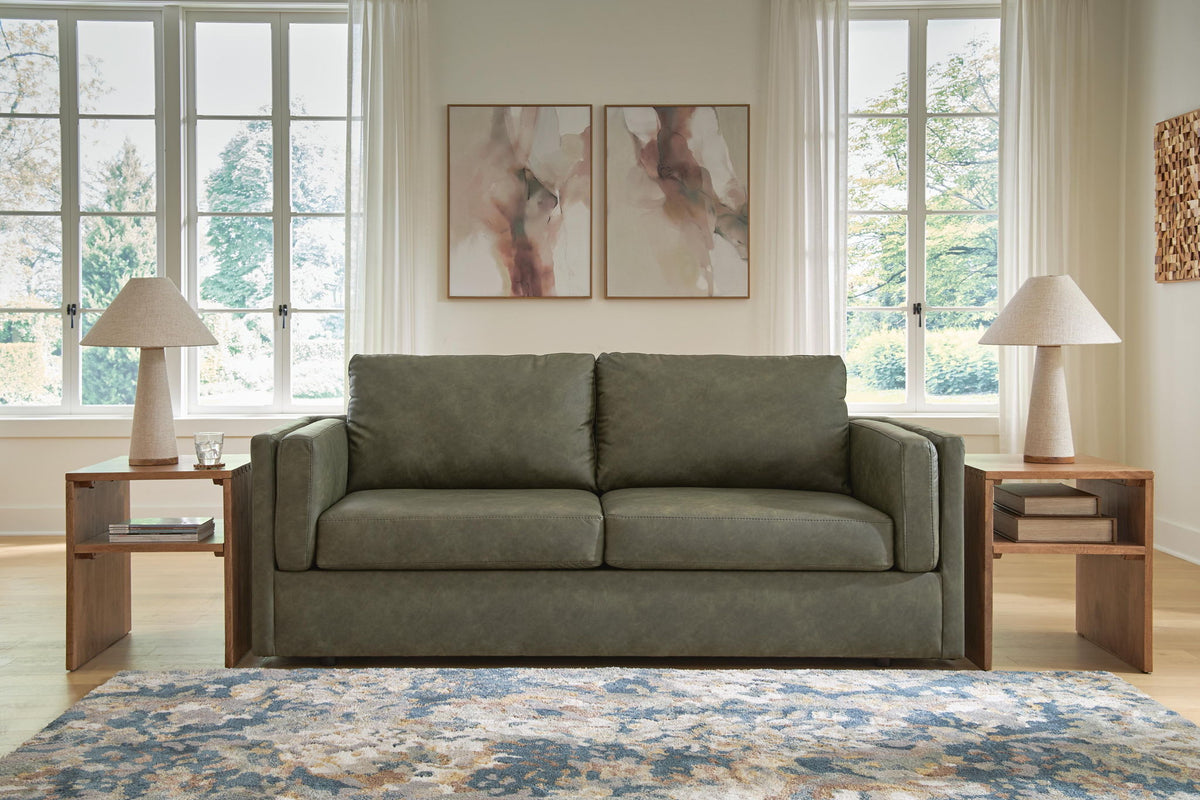 A two-seater sofa upholstered in muted sage green fabric, styled in a bright living room with two wooden side tables, table lamps, a patterned rug, and abstract wall art.