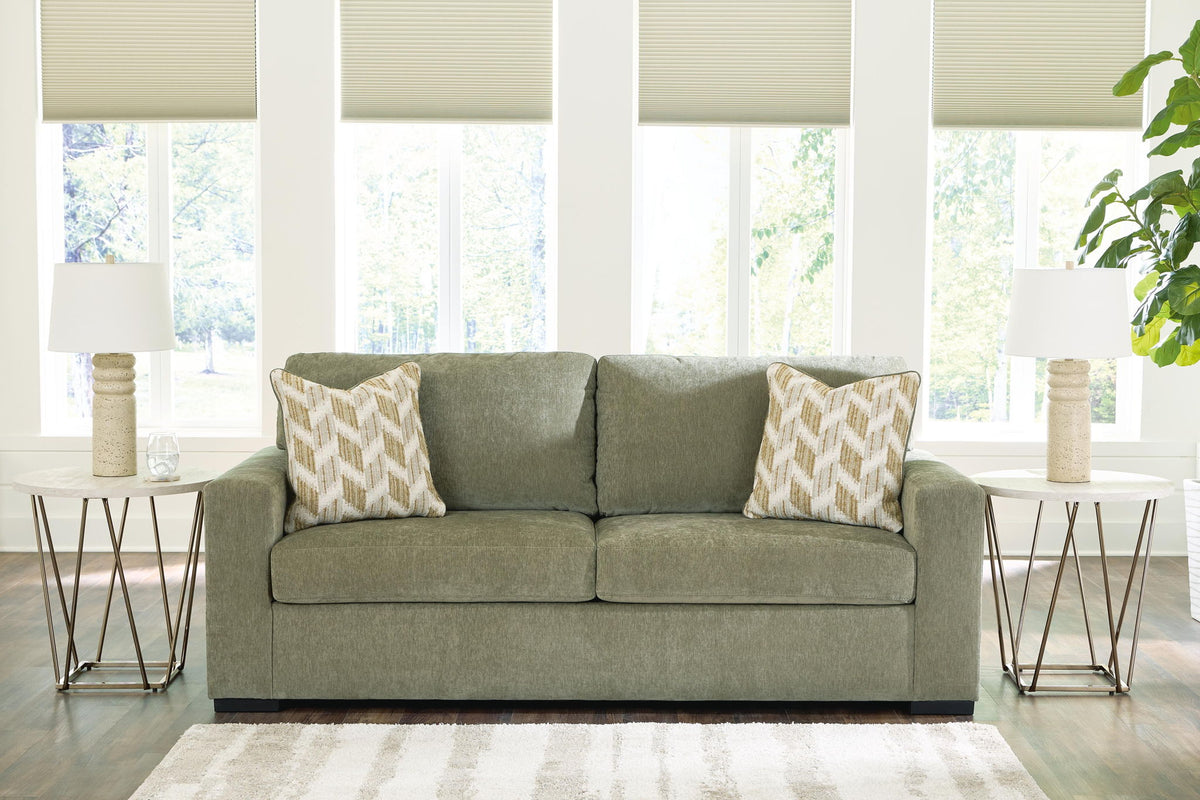 An olive green two-seater loveseat sofa with two patterned beige and white accent pillows, styled in a bright living room with side tables, lamps, and windows.