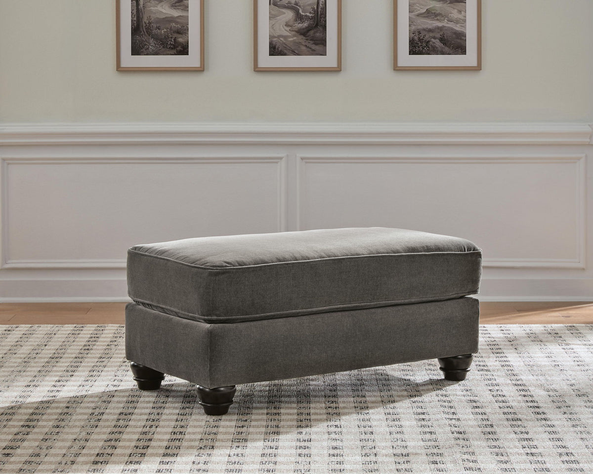 A gray upholstered ottoman with dark turned legs, shown in a styled room setting on a patterned rug against a white wainscoted wall.