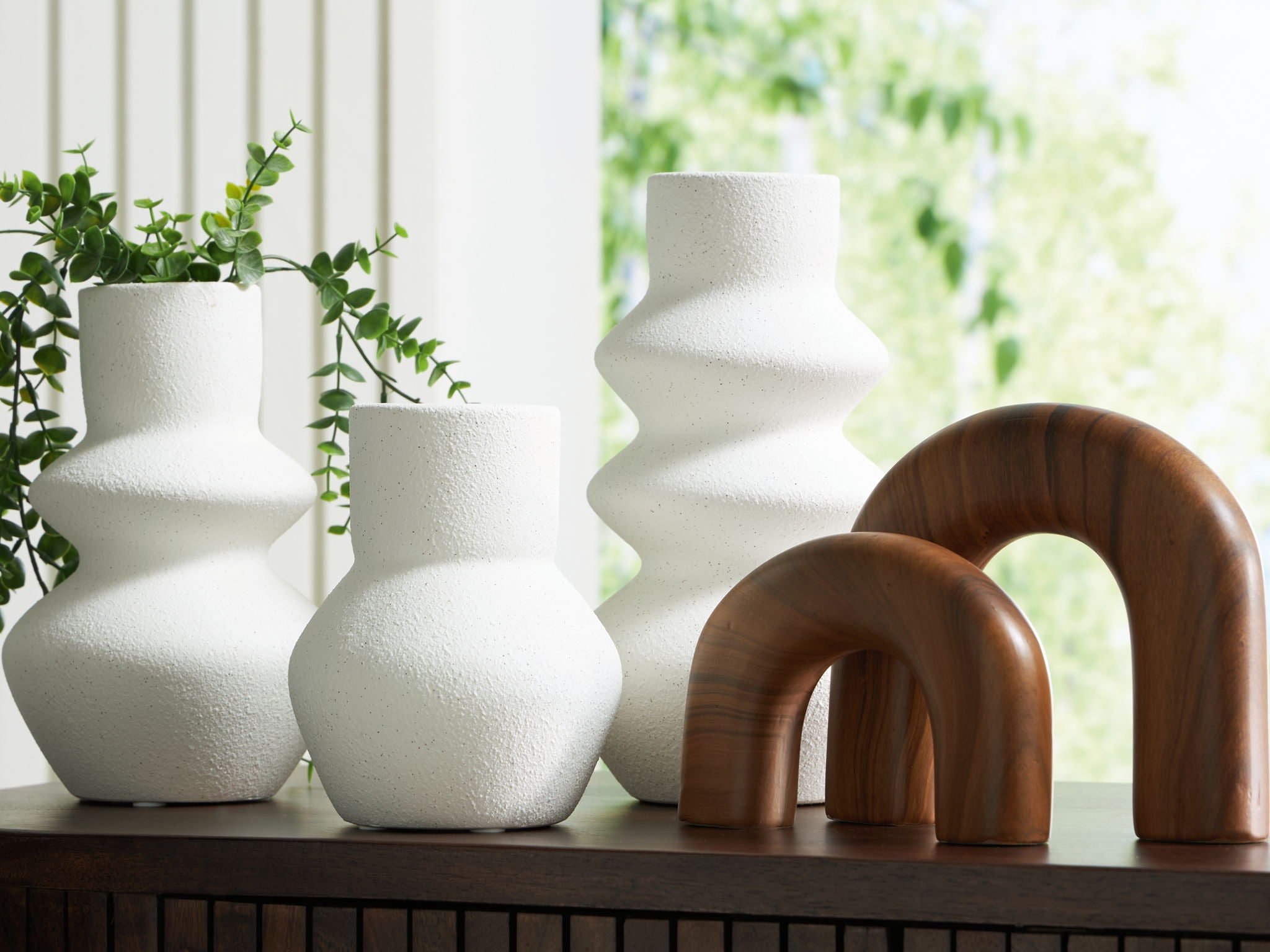 White textured vases and a brown arch-shaped object on a table with a blurred green outdoor background.