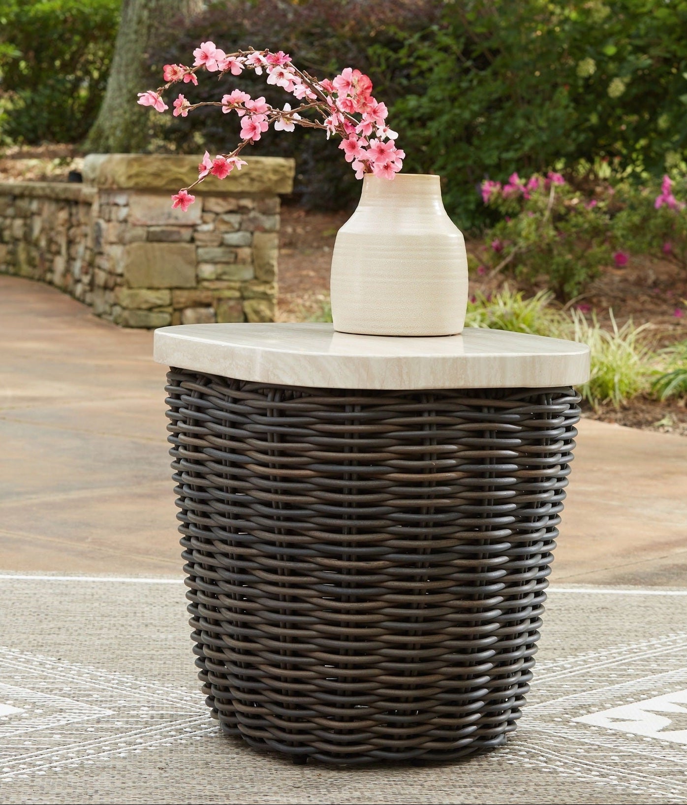 Wicker side table with a white vase and pink flowers on a patio