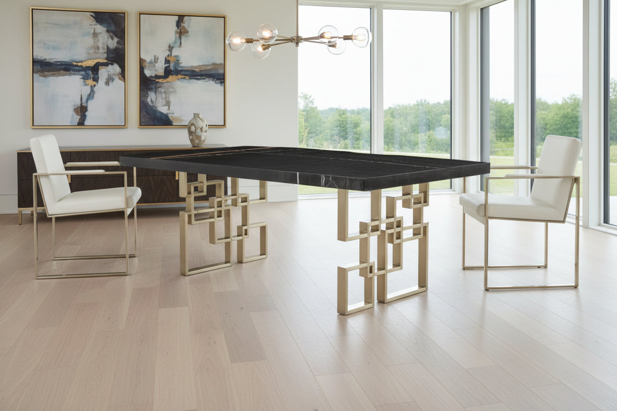 Dining table with geometric brass legs and two chairs in a room scene.