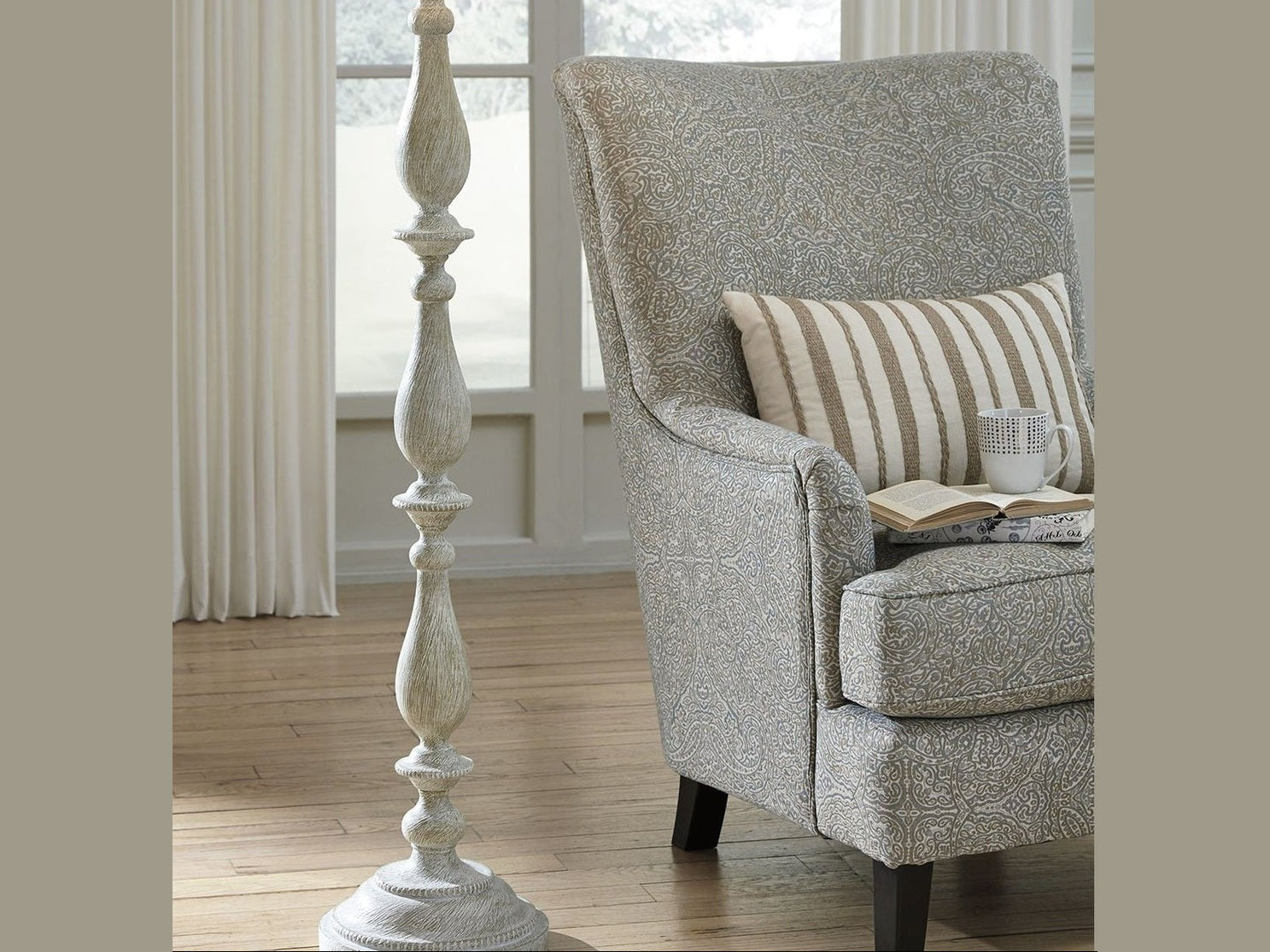 Gray armchair with a striped pillow and a book in a room with a large window and decorative lamp.