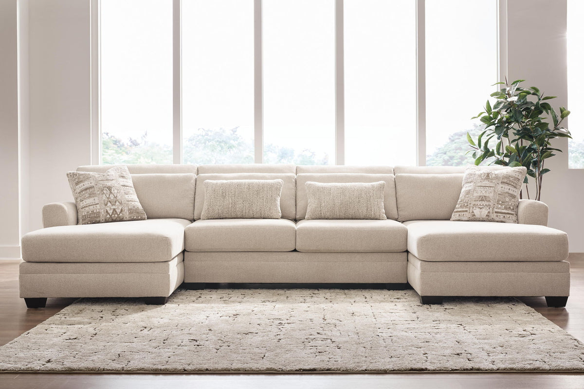 A light beige upholstered sectional sofa with a right-facing chaise and four matching throw pillows, styled in a bright living room with a textured rug, large windows, and a potted plant.