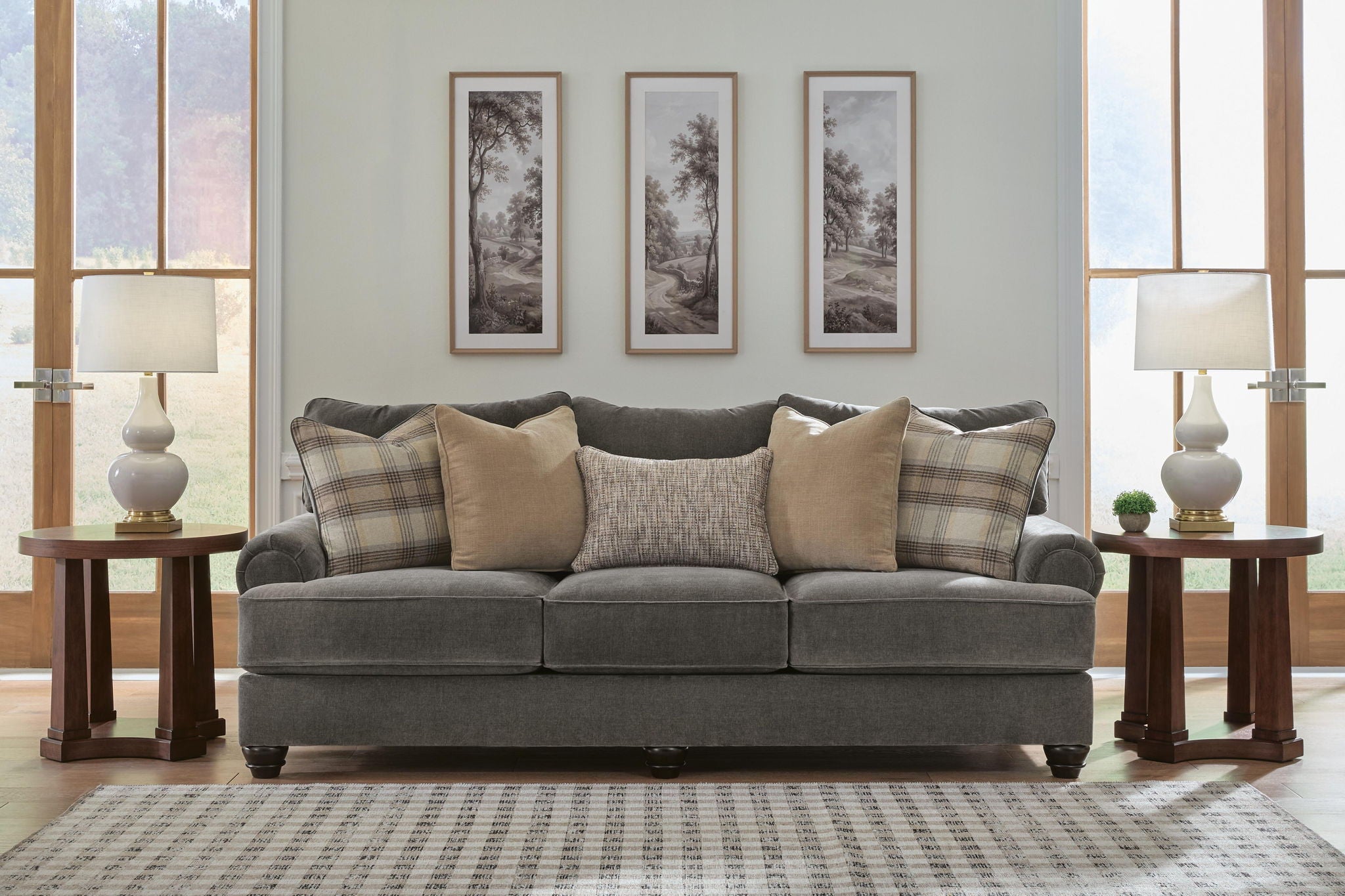 A gray upholstered loveseat with beige and plaid throw pillows, shown in a staged living room setting with two wooden side tables, table lamps, wall art, and a patterned rug.