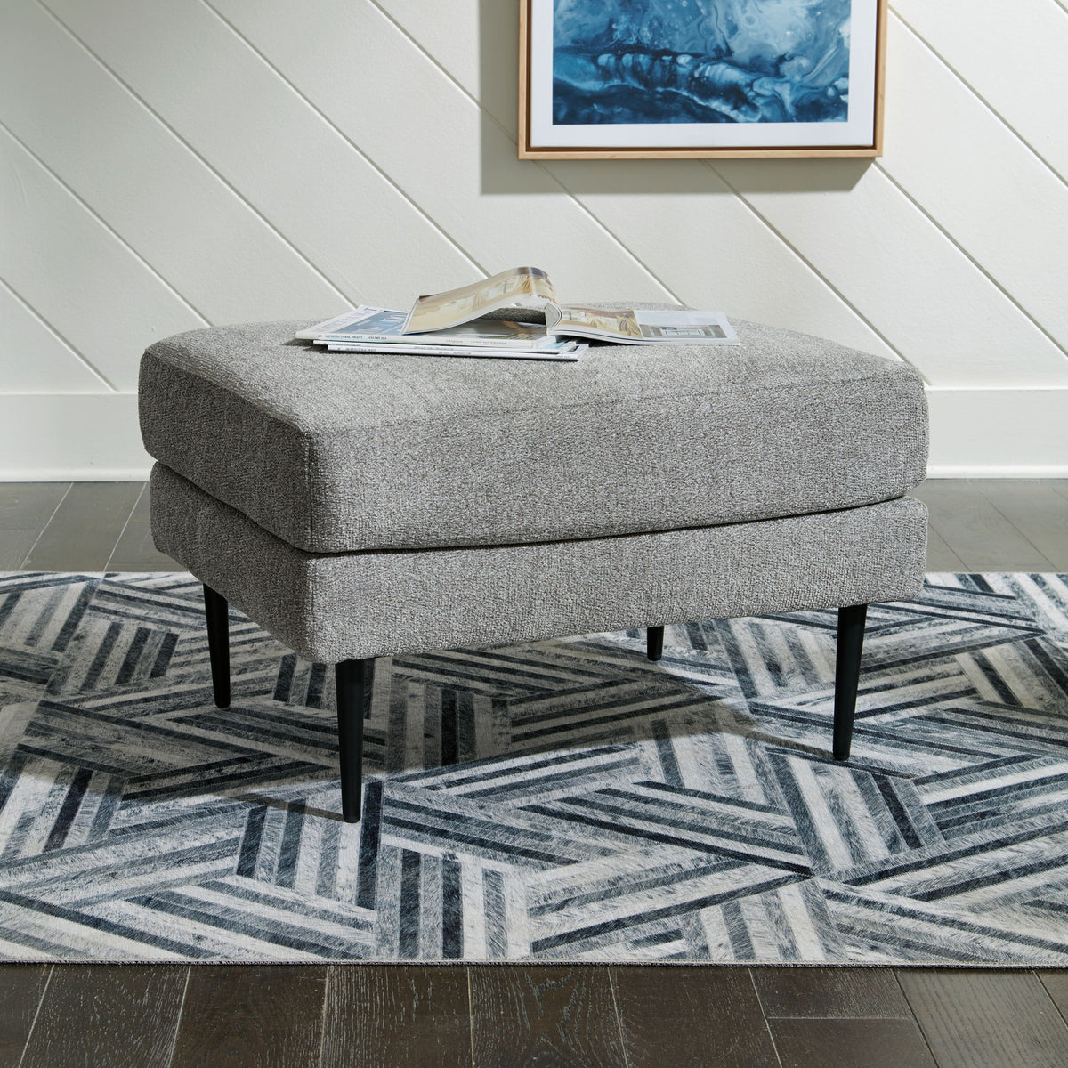 Gray upholstered ottoman on a geometric patterned rug with a framed picture on the wall.