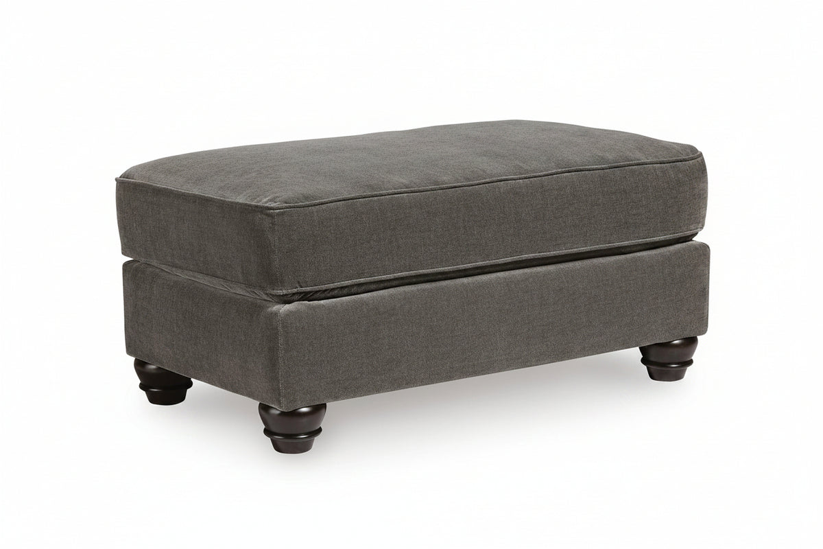 A dark gray upholstered ottoman with a thick cushioned top and turned dark wooden feet, shown on a white background.