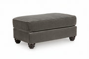 A dark gray upholstered ottoman with a thick cushioned top and turned dark wooden feet, shown on a white background.