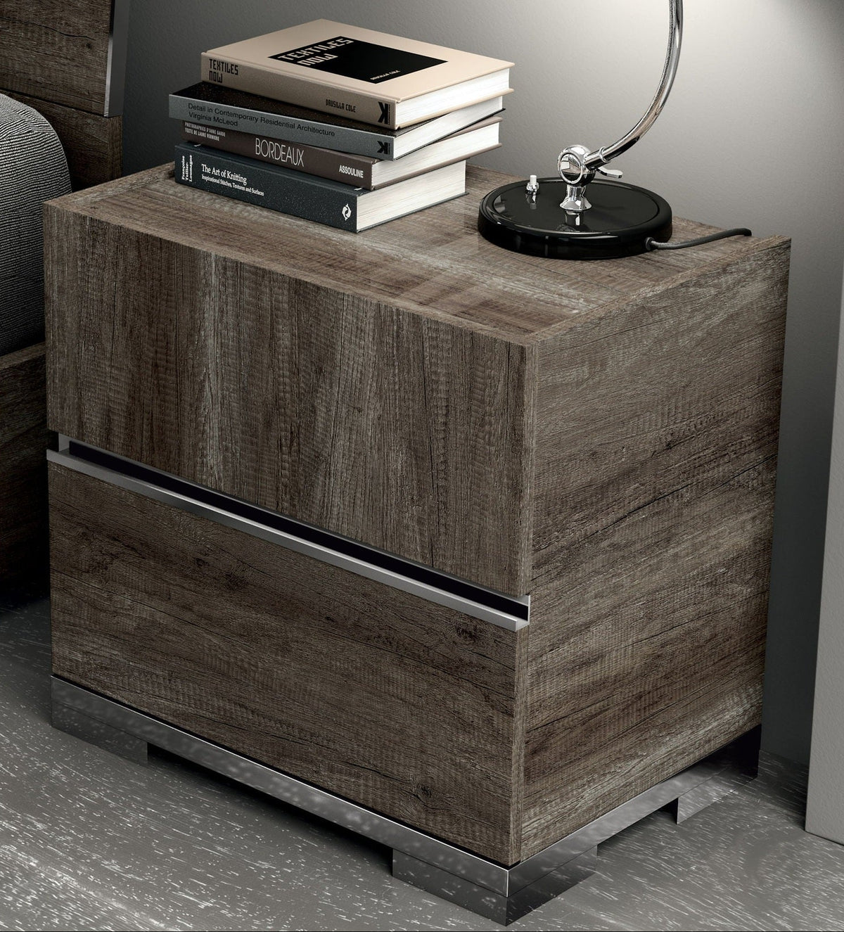 A modern two-drawer nightstand with a brown wood grain finish and a slim metallic handle, styled with books and a lamp on top.