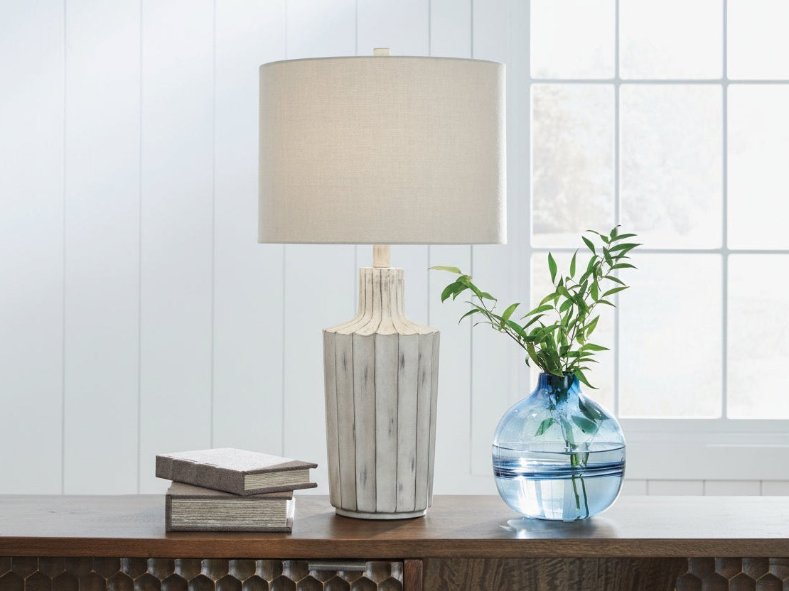 The Imre table lamp with a beige shade and decorative vase on a wooden surface against a white paneled wall.