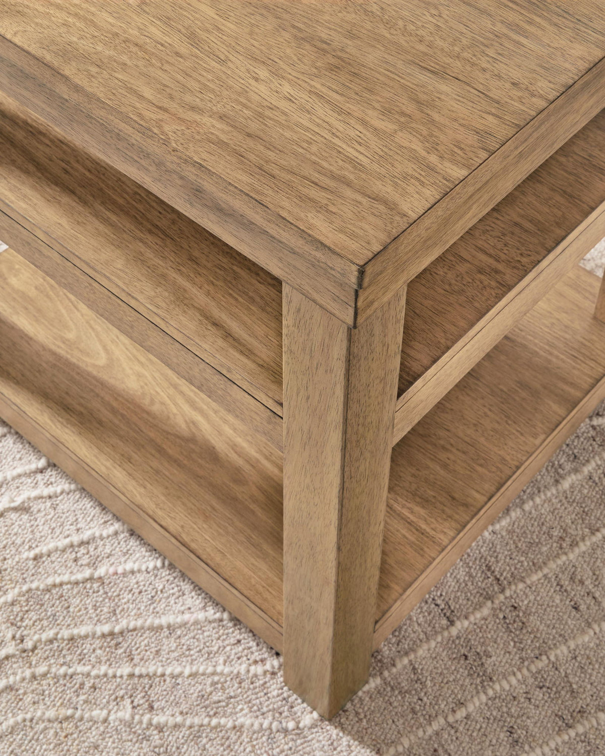Close-up of a wooden side table or coffee table corner with a warm medium-brown wood grain finish, shown resting on a light beige textured rug.