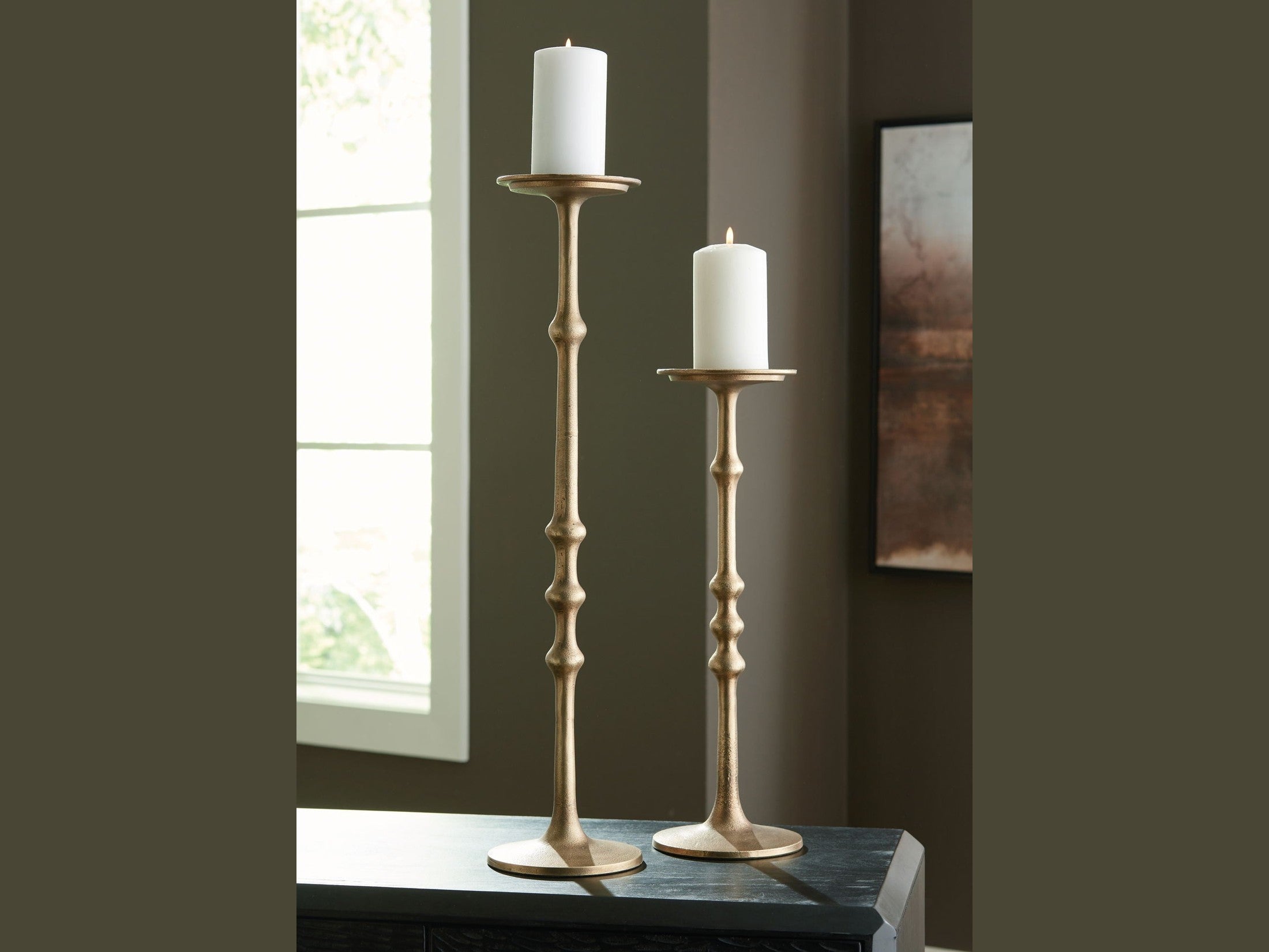 Two gold candle holders with white candles on a dark surface near a window.