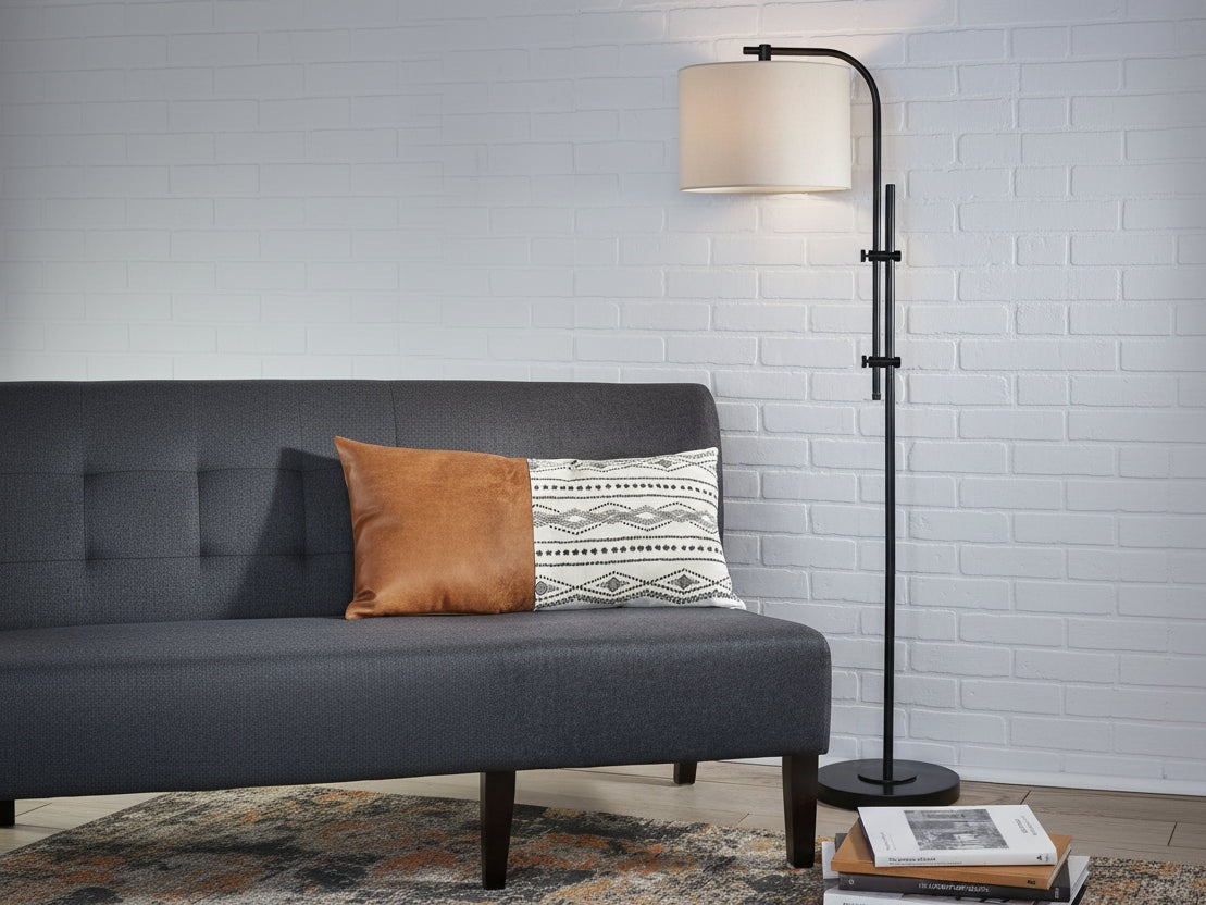 Baronvale gray sofa with a patterned pillow and a standing lamp against a white brick wall.