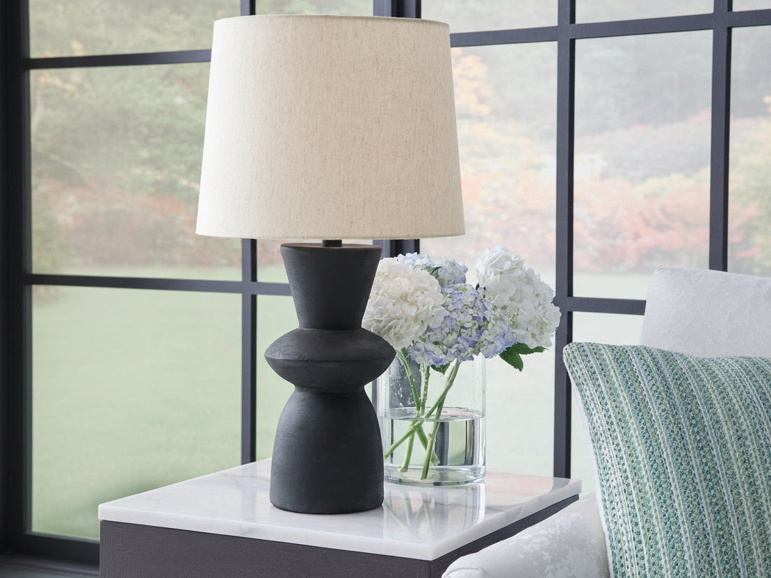 Scarbot table lamp with a beige shade on a table next to a vase of flowers, with a blurred window in the background.