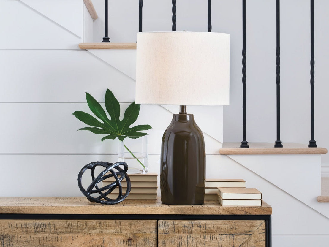 The Jyler table lamp with a white shade is on a wooden surface next to books and a plant.