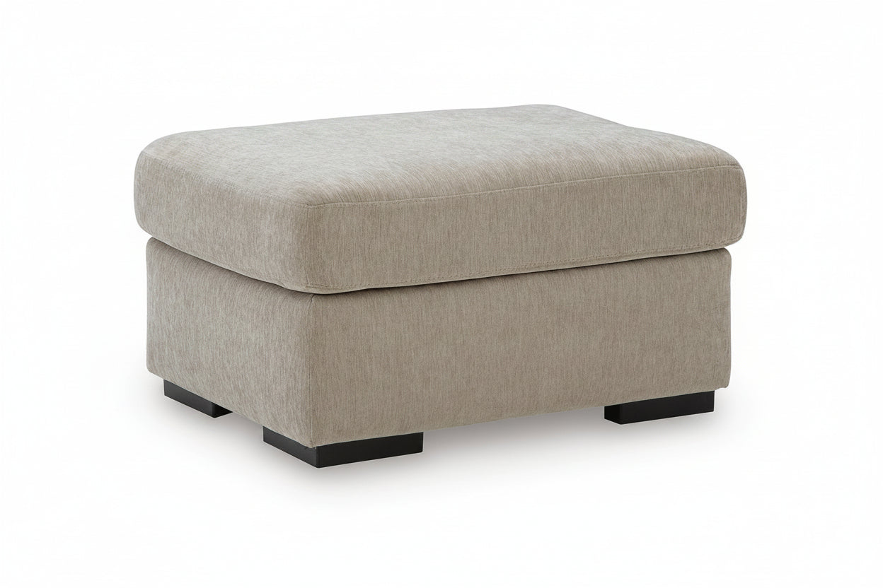 A square upholstered ottoman with a thick cushioned top and a low boxy base, shown on a plain white background. The fabric is a light warm gray or taupe, and the ottoman sits on short dark block feet.