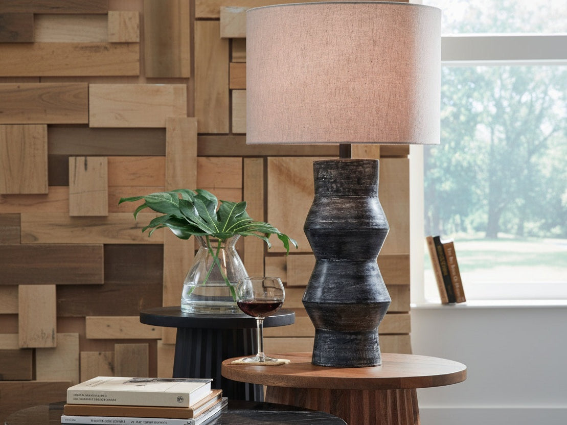Kerbert table lamp with a textured base and beige shade on a wooden table, with a glass of red wine and a plant in the background.