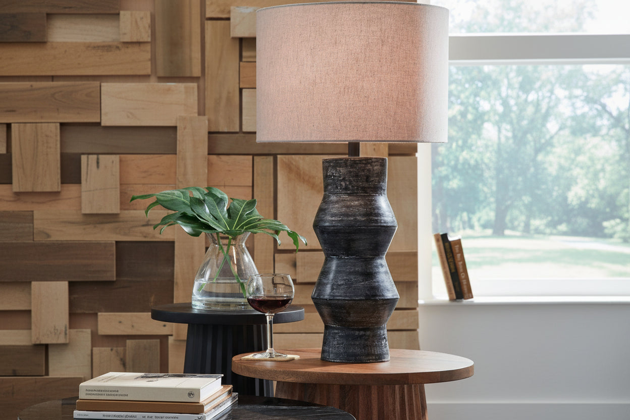 Kerbert table lamp with a textured base and beige shade on a wooden table, with a glass of red wine and a plant in the background.