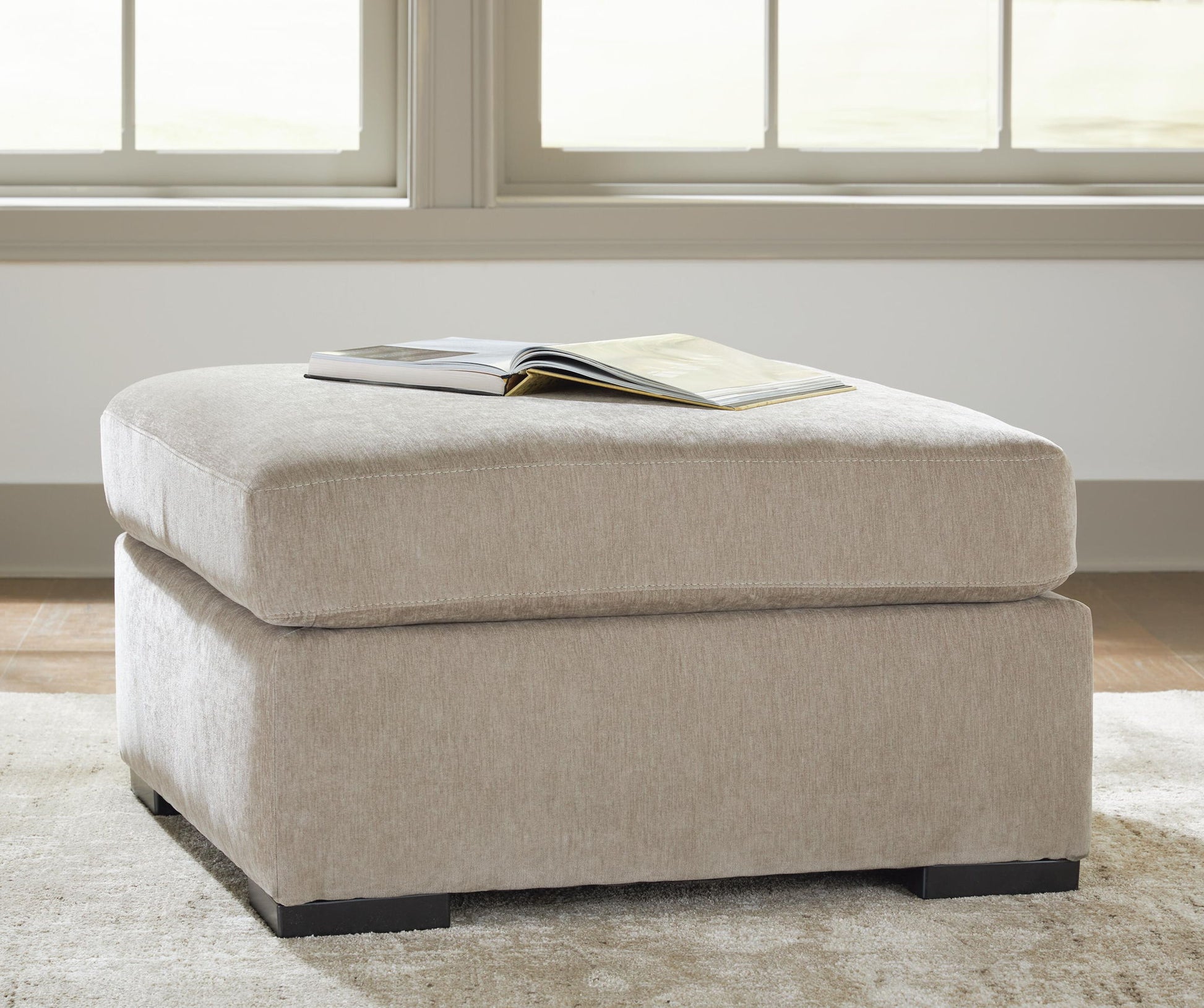 A two-tier upholstered ottoman in a light beige textured fabric, styled with an open book on top, set on a neutral rug in front of a bright window.