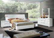 Asti modern bedroom with white furniture, including a bed, nightstands, and dresser, in a room with large windows showing greenery.