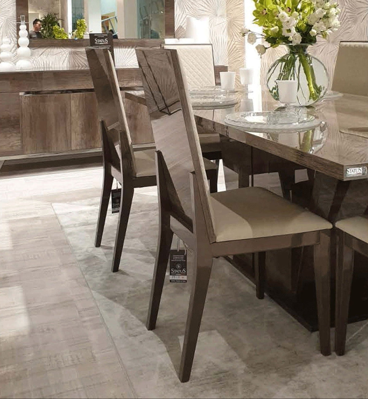 A dining room scene showing a dark wood dining table with upholstered chairs, a glass vase of flowers on the table, and a mirrored sideboard in the background.