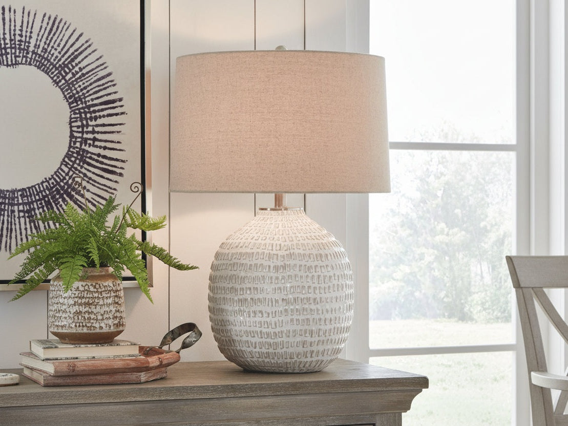 Jamon table lamp with textured base and beige shade on a wooden surface near a window.