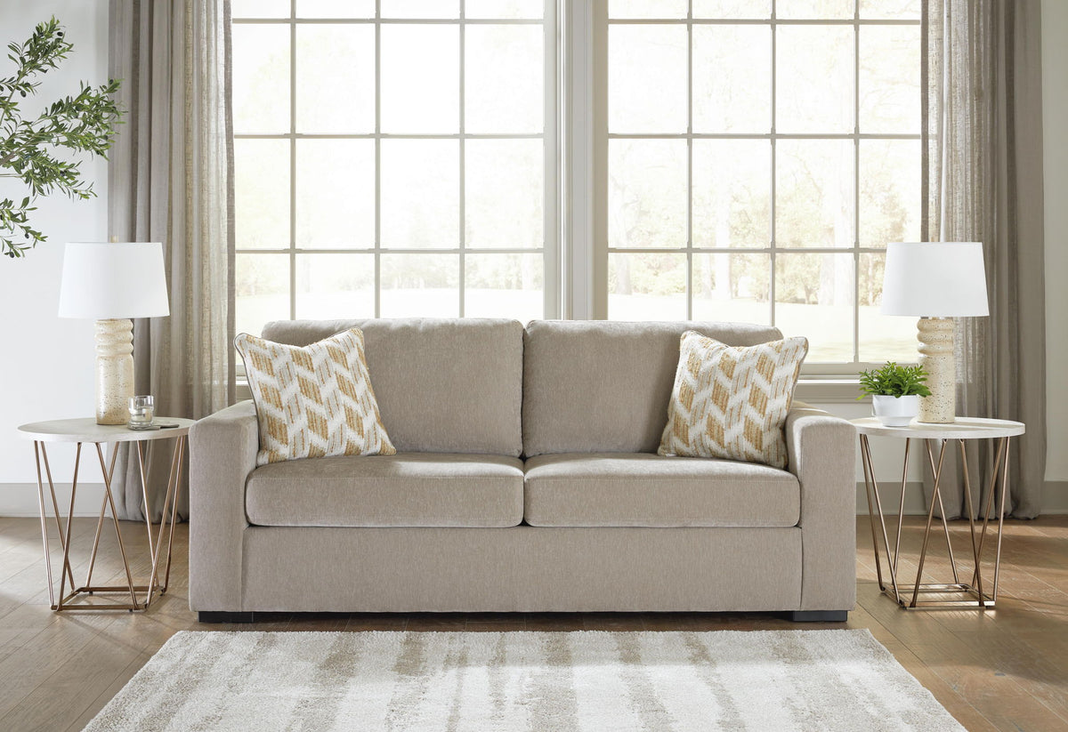 A light beige two-seater sofa with two patterned throw pillows, styled in a bright living room with two side tables, lamps, and large windows.